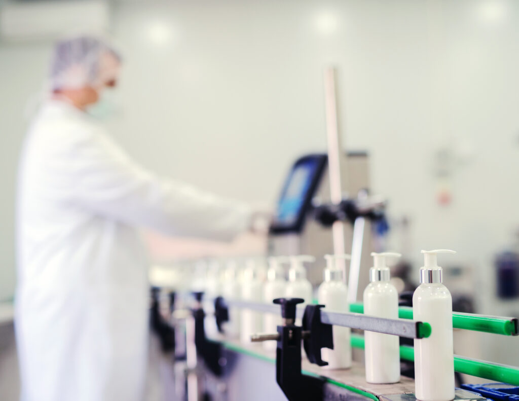 Picture of lotion bottles on production line. Bottles of cosmetic products in factory production line. Blurred picture of man using control panel in background.